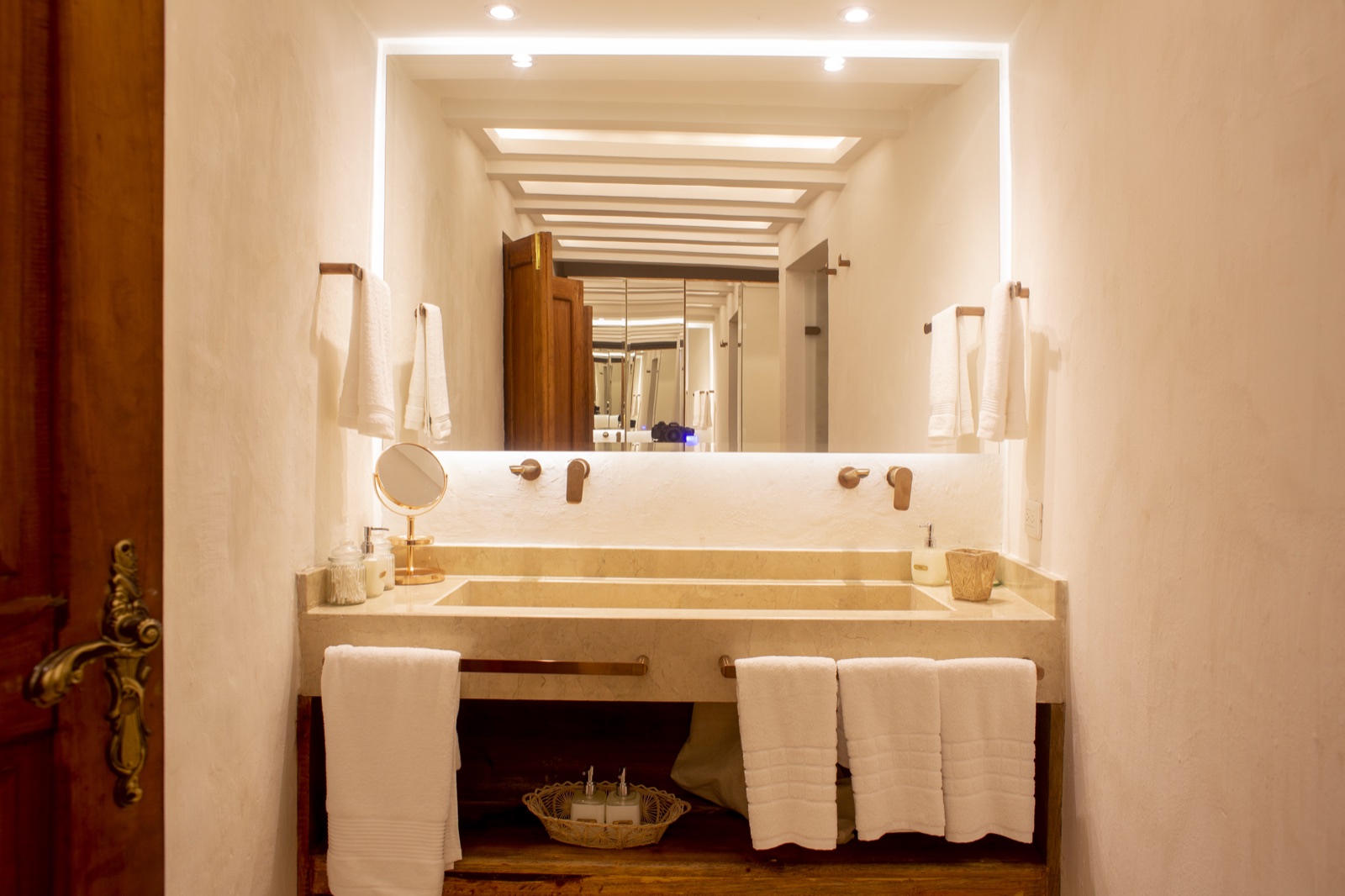 Casa Palmas en-suite bathroom with backlit floating mirror, dual bronze wall-mount faucets, and travertine double sink